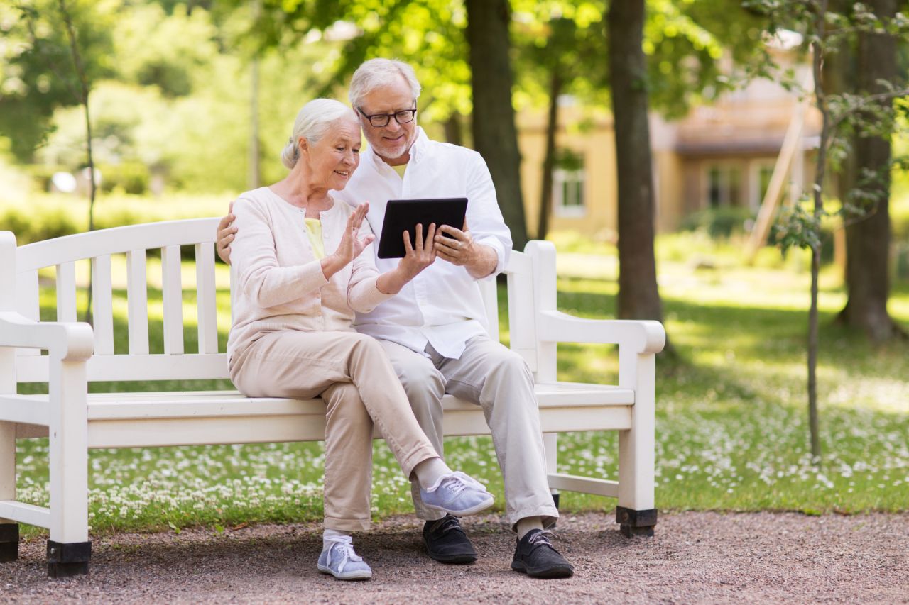Couple âgé assis sur un banc regardant une tablette, dans un parc verdoyant.